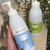 Hand holding a blue-labelled MooGoo Flake Free shampoo pump bottle, with the green-labelled conditioner and trailing foliage behind.