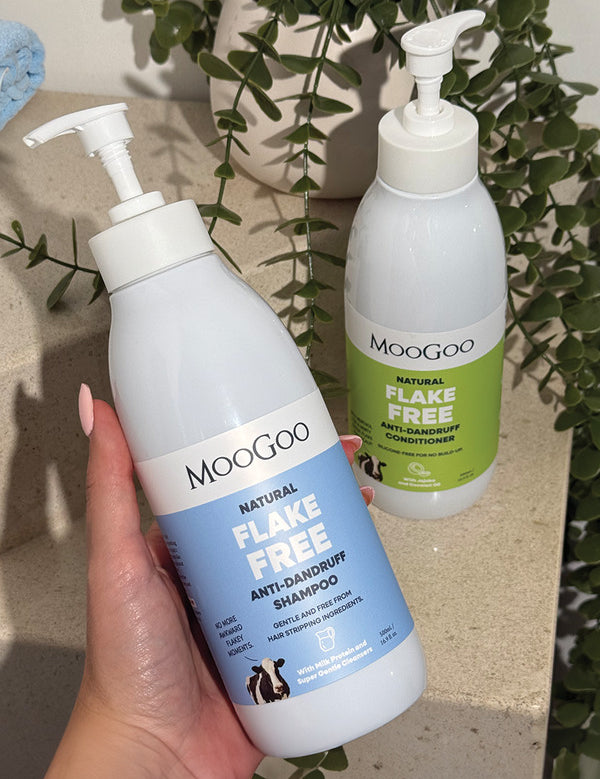 Hand holding a blue-labelled MooGoo Flake Free shampoo pump bottle, with the green-labelled conditioner and trailing foliage behind.