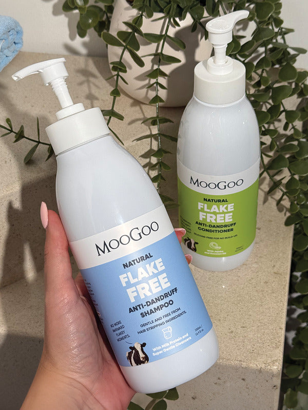 Hand holding a blue-labelled MooGoo Flake Free shampoo pump bottle, with the green-labelled conditioner and trailing foliage behind.