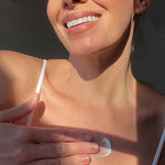 Smiling person in a white camisole rubbing a dab of white moisturiser onto their upper chest in bright sunlight.