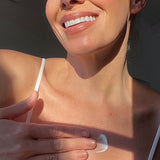 Smiling person in a white camisole rubbing a dab of white moisturiser onto their upper chest in bright sunlight.