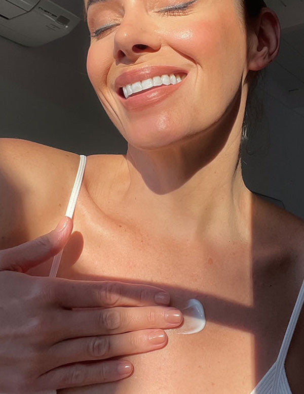 Smiling person in a white camisole rubbing a dab of white moisturiser onto their upper chest in bright sunlight.