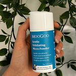Hand holding a blue-labelled MooGoo Gentle Exfoliating Micro Powder container with a white cap against trailing green foliage.