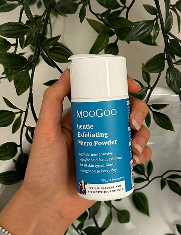 Hand holding a blue-labelled MooGoo Gentle Exfoliating Micro Powder container with a white cap against trailing green foliage.