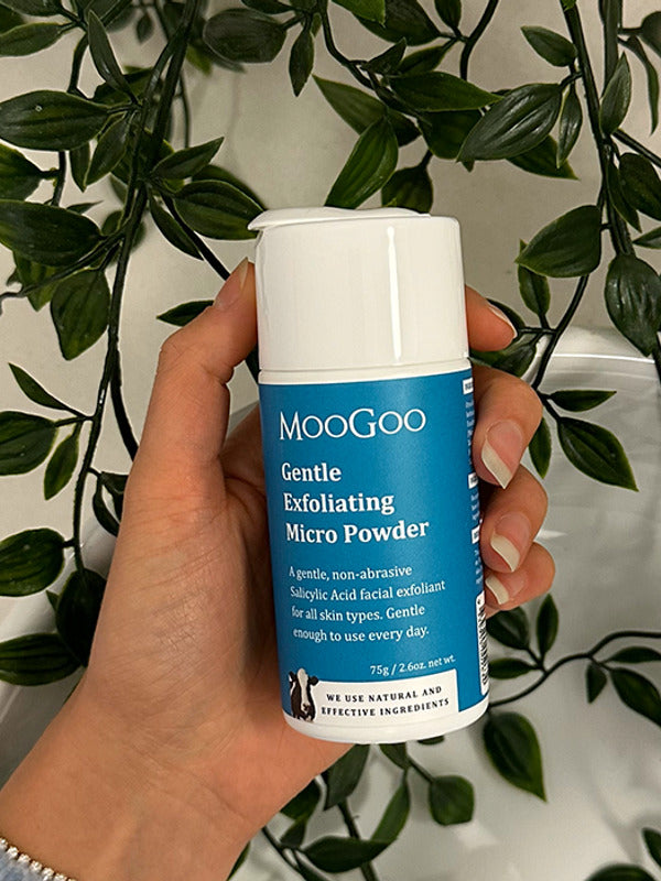 Hand holding a blue-labelled MooGoo Gentle Exfoliating Micro Powder container with a white cap against trailing green foliage.