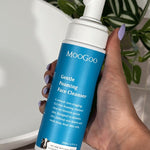 Hand holding a MooGoo Gentle Foaming Face Cleanser 200ml pump bottle beside a white basin and green leaves.