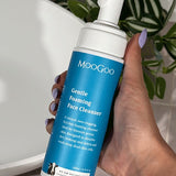 Hand holding a MooGoo Gentle Foaming Face Cleanser 200ml pump bottle beside a white basin and green leaves.
