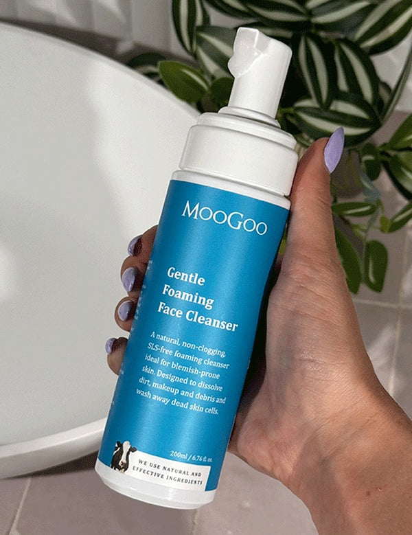 Hand holding a MooGoo Gentle Foaming Face Cleanser 200ml pump bottle beside a white basin and green leaves.