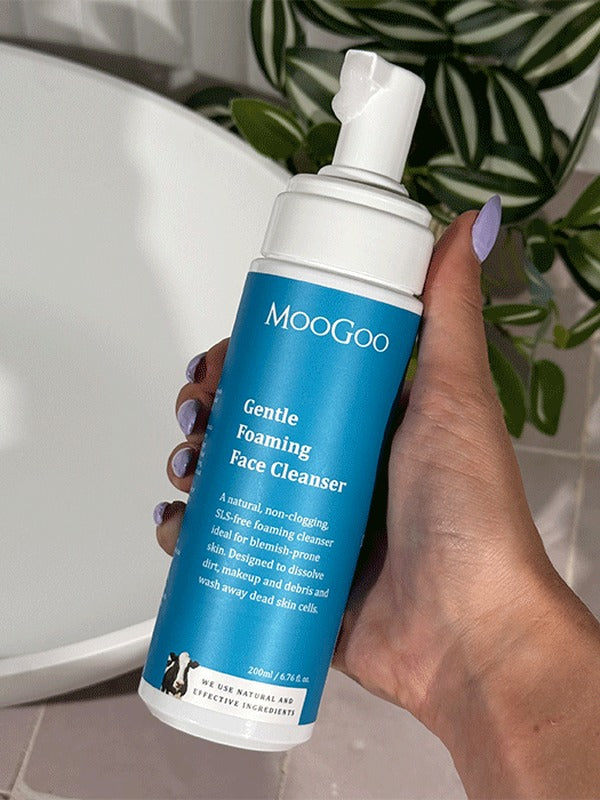 Hand holding a MooGoo Gentle Foaming Face Cleanser 200ml pump bottle beside a white basin and green leaves.