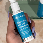 Hand with lilac nail polish holding a blue-labelled MooGoo Hyaluronic Acid and Niacinamide Active Serum pump bottle, with another bottle on a green tray behind.