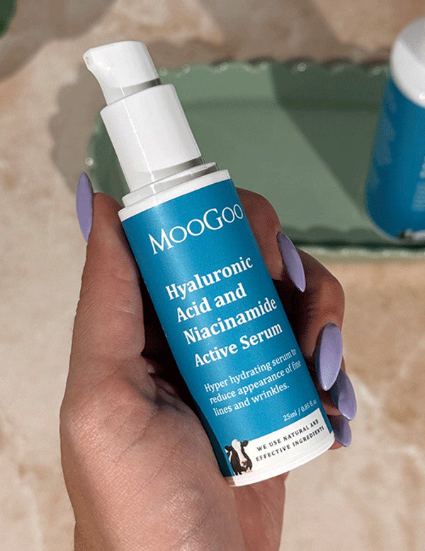 Hand with lilac nail polish holding a blue-labelled MooGoo Hyaluronic Acid and Niacinamide Active Serum pump bottle, with another bottle on a green tray behind.