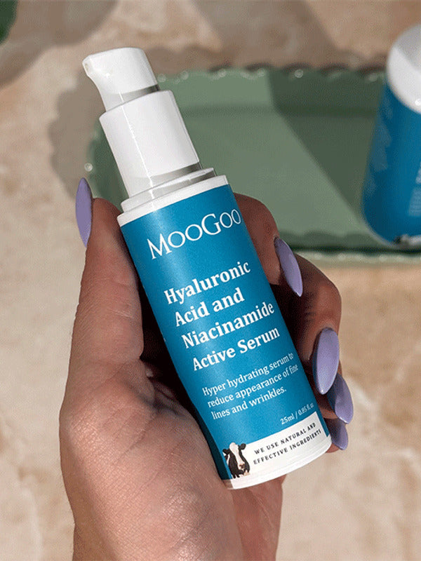 Hand with lilac nail polish holding a blue-labelled MooGoo Hyaluronic Acid and Niacinamide Active Serum pump bottle, with another bottle on a green tray behind.
