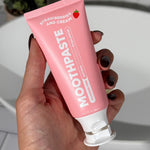 Hand holding an open pink MooGoo Kids Strawberries and Cream Moothpaste tube beside a white bathroom basin.