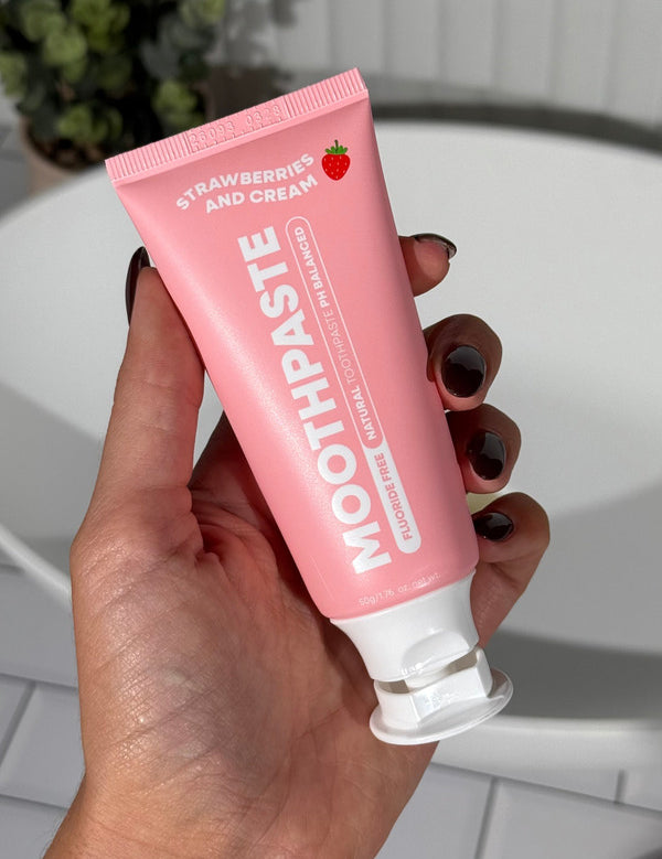 Hand holding an open pink MooGoo Kids Strawberries and Cream Moothpaste tube beside a white bathroom basin.