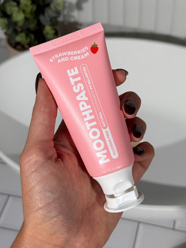Hand holding an open pink MooGoo Kids Strawberries and Cream Moothpaste tube beside a white bathroom basin.