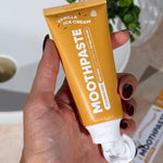 Hand holding an open MooGoo Kids Vanilla Ice Cream Moothpaste tube beside a basin and matching carton.