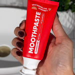 Hand holding an open red MooGoo Kids Watermelon Sherbet Moothpaste tube beside a bathroom basin.