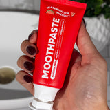 Hand holding an open red MooGoo Kids Watermelon Sherbet Moothpaste tube beside a bathroom basin.