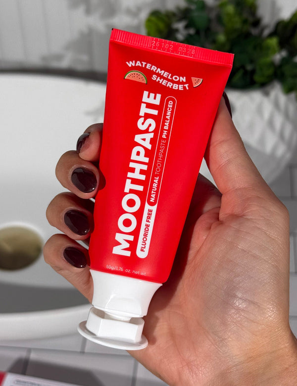 Hand holding an open red MooGoo Kids Watermelon Sherbet Moothpaste tube beside a bathroom basin.