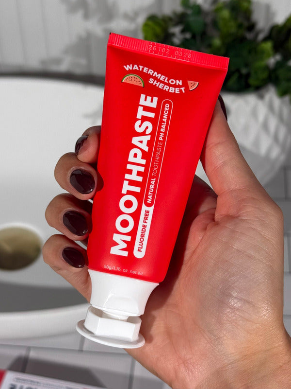Hand holding an open red MooGoo Kids Watermelon Sherbet Moothpaste tube beside a bathroom basin.