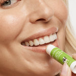Close-up of a smiling person applying MooGoo Cow Lick lip balm to their lower lip from a green-and-white tube.