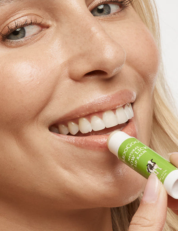 Close-up of a smiling person applying MooGoo Cow Lick lip balm to their lower lip from a green-and-white tube.