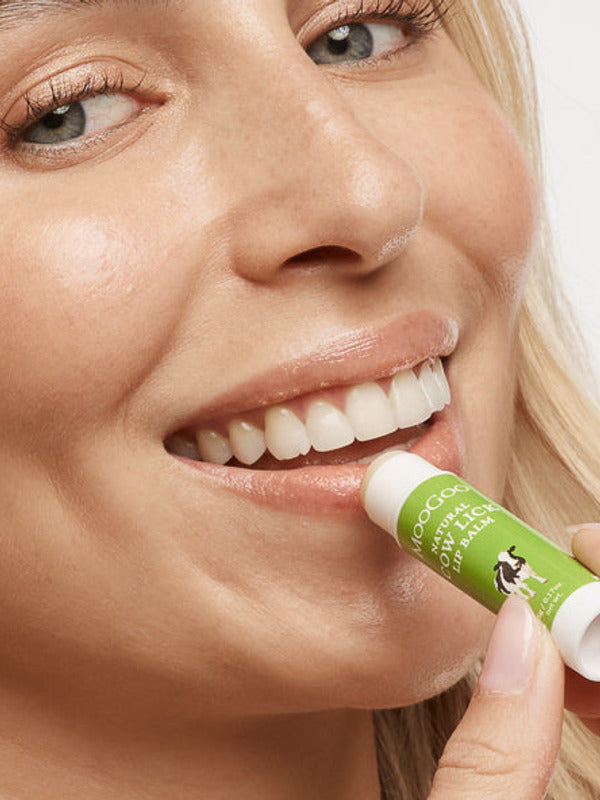 Close-up of a smiling person applying MooGoo Cow Lick lip balm to their lower lip from a green-and-white tube.