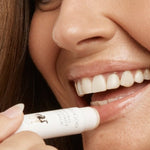 Close-up lifestyle photograph of a smiling woman applying MooGoo Natural Coconut Lip Balm to her lower lip from the white product tube.