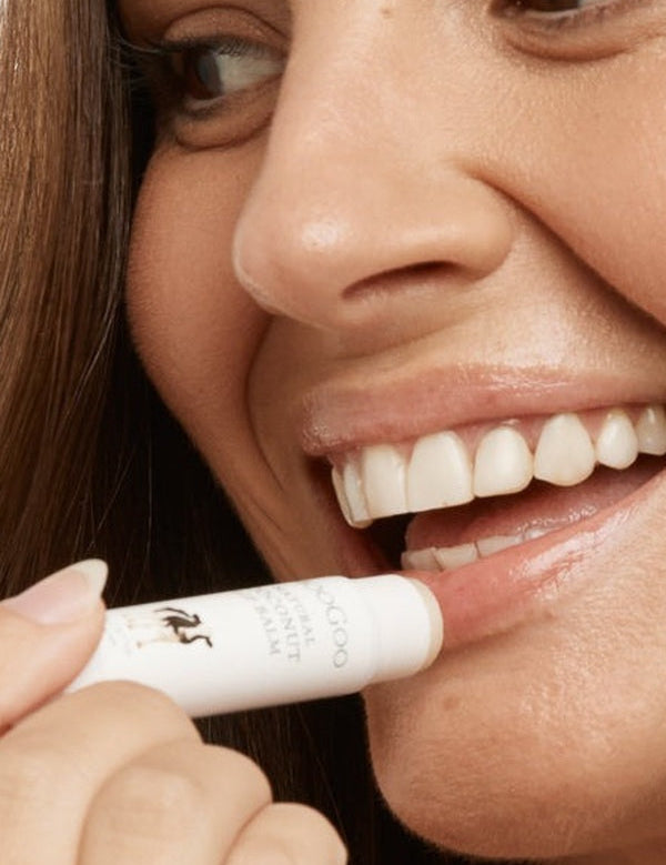 Close-up lifestyle photograph of a smiling woman applying MooGoo Natural Coconut Lip Balm to her lower lip from the white product tube.