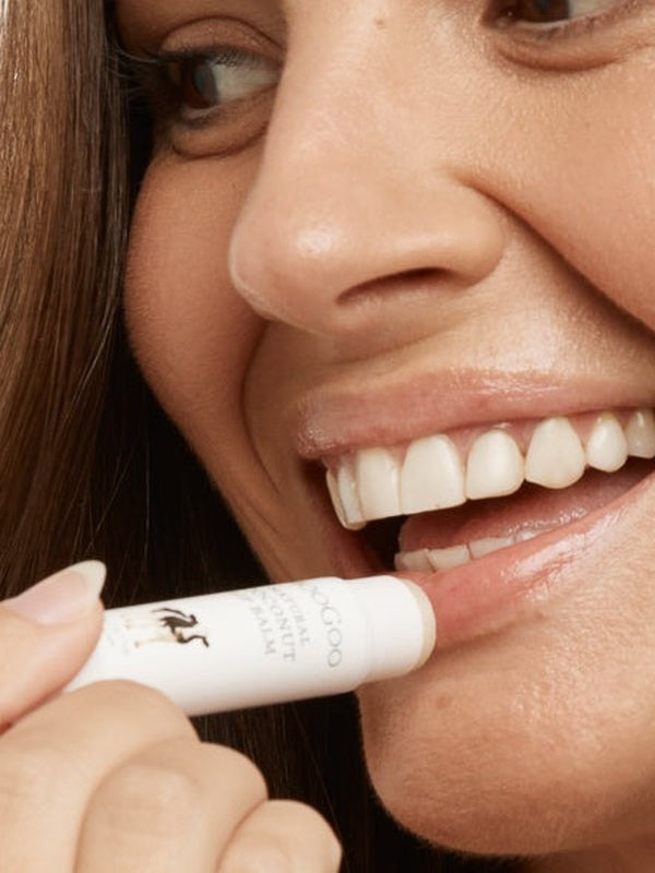 Close-up lifestyle photograph of a smiling woman applying MooGoo Natural Coconut Lip Balm to her lower lip from the white product tube.