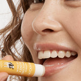 Close-up of a smiling person applying MooGoo Tingling Honey lip balm to their lower lip from a yellow-and-white tube.