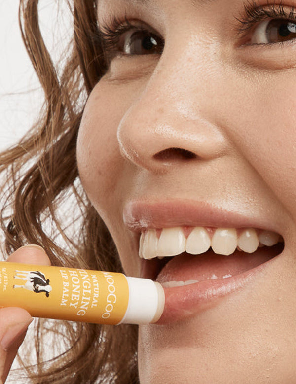 Close-up of a smiling person applying MooGoo Tingling Honey lip balm to their lower lip from a yellow-and-white tube.