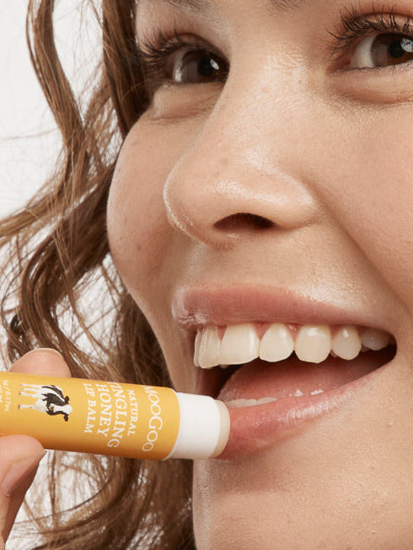 Close-up of a smiling person applying MooGoo Tingling Honey lip balm to their lower lip from a yellow-and-white tube.
