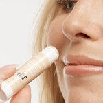 Close-up of a person holding an uncapped peach-and-white MooGoo Vanilla Dream lip balm tube beside their upper lip.