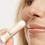 Close-up of a person holding an uncapped peach-and-white MooGoo Vanilla Dream lip balm tube beside their upper lip.