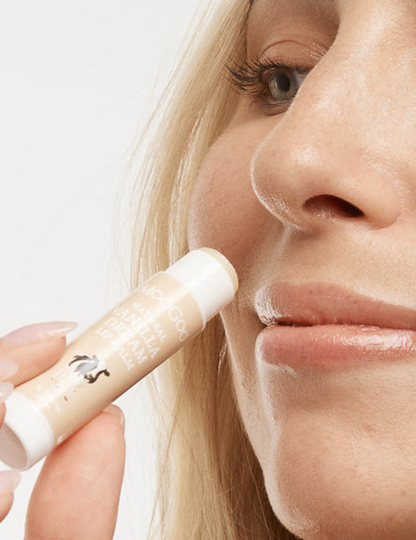 Close-up of a person holding an uncapped peach-and-white MooGoo Vanilla Dream lip balm tube beside their upper lip.