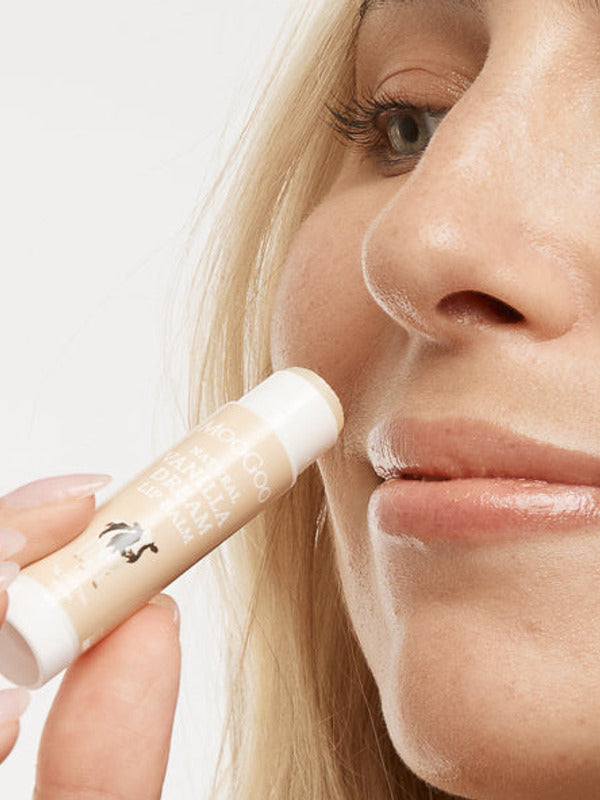 Close-up of a person holding an uncapped peach-and-white MooGoo Vanilla Dream lip balm tube beside their upper lip.
