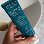 Hand holding a dark turquoise MooGoo Baby and Child Eczema and Psoriasis Cream tube beside a white basin with a brass-coloured plug.