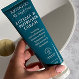 Hand holding a dark turquoise MooGoo Baby and Child Eczema and Psoriasis Cream tube beside a white basin with a brass-coloured plug.