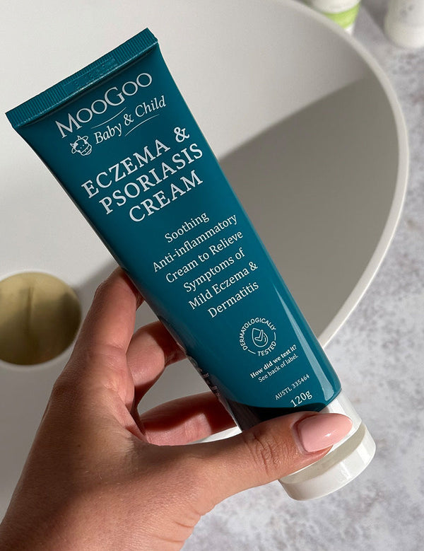 Hand holding a dark turquoise MooGoo Baby and Child Eczema and Psoriasis Cream tube beside a white basin with a brass-coloured plug.