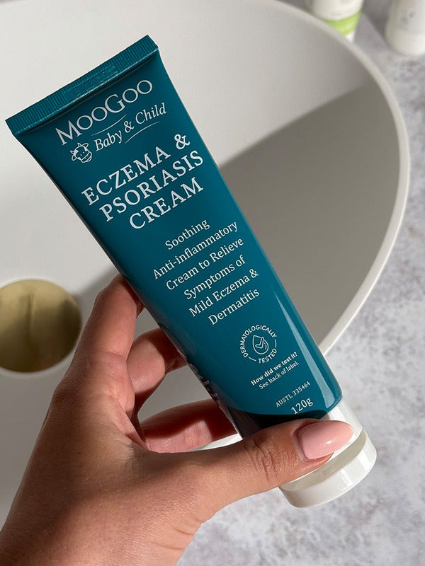 Hand holding a dark turquoise MooGoo Baby and Child Eczema and Psoriasis Cream tube beside a white basin with a brass-coloured plug.