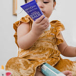 Young child in a mustard-coloured outfit sitting among toiletries and looking at a purple MooGoo Nipple Balm tube held close to their face.