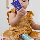 Young child in a mustard-coloured outfit sitting among toiletries and looking at a purple MooGoo Nipple Balm tube held close to their face.