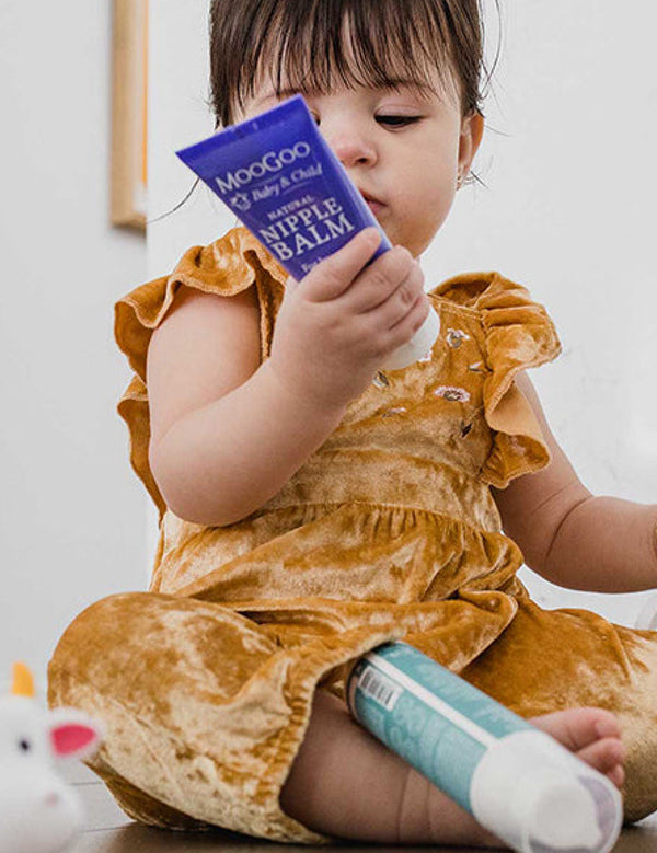 Young child in a mustard-coloured outfit sitting among toiletries and looking at a purple MooGoo Nipple Balm tube held close to their face.