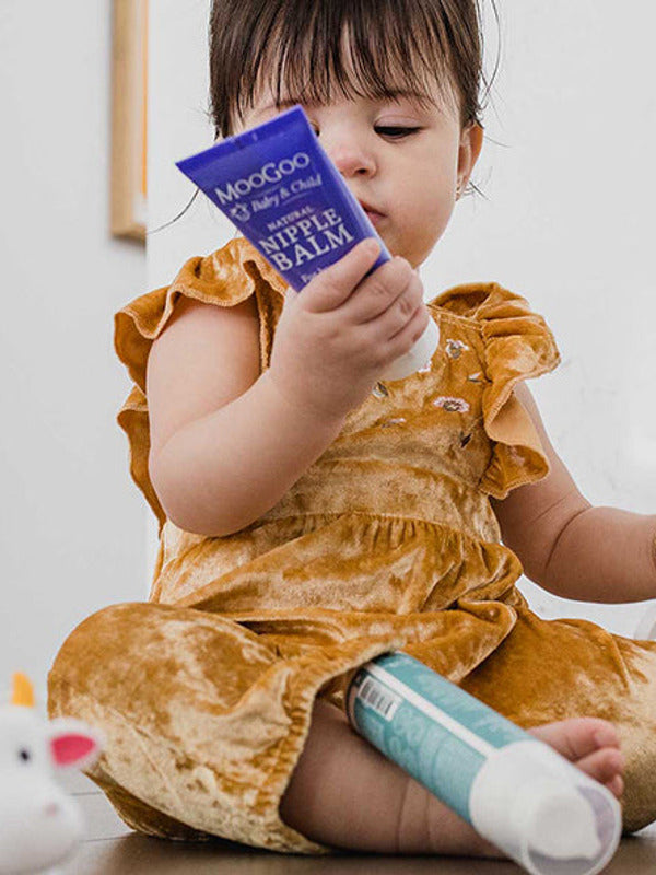 Young child in a mustard-coloured outfit sitting among toiletries and looking at a purple MooGoo Nipple Balm tube held close to their face.