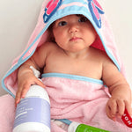 Baby wrapped in a pink-and-blue hooded towel with butterfly wings on the hood, with a purple-labelled wash bottle and green MooGoo moisturiser tube in their lap.
