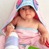 Baby wrapped in a pink-and-blue hooded towel with butterfly wings on the hood, with a purple-labelled wash bottle and green MooGoo moisturiser tube in their lap.