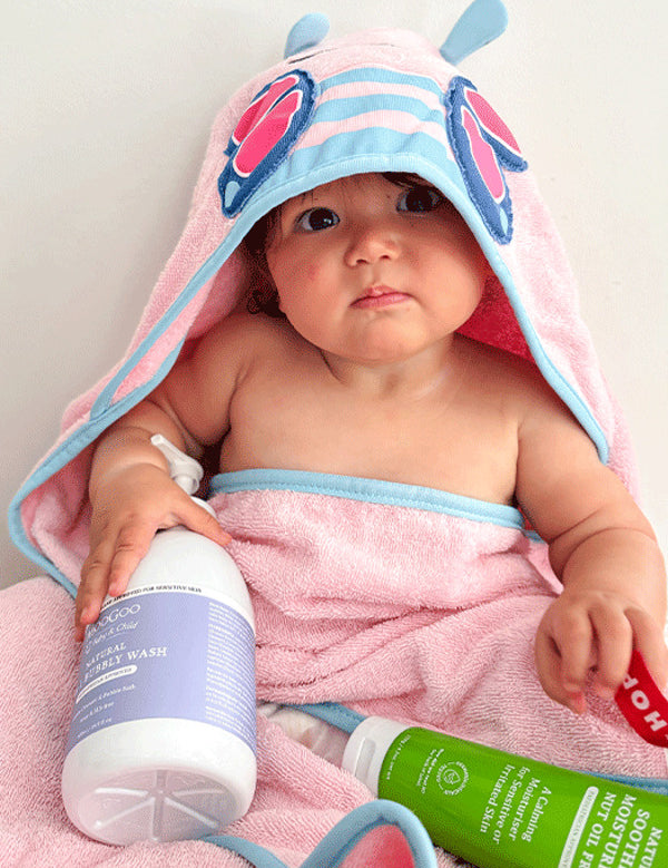 Baby wrapped in a pink-and-blue hooded towel with butterfly wings on the hood, with a purple-labelled wash bottle and green MooGoo moisturiser tube in their lap.