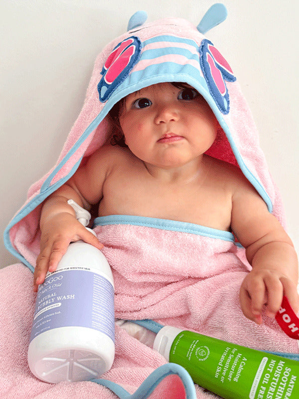 Baby wrapped in a pink-and-blue hooded towel with butterfly wings on the hood, with a purple-labelled wash bottle and green MooGoo moisturiser tube in their lap.