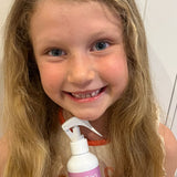 Smiling child with long wavy blonde hair holding a pink-labelled MooGoo detangling spray bottle with a white trigger nozzle.